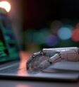 Robotic Hand Interacting with Laptop Displaying Code. A close-up shot of a robotic hand resting on a laptop keyboard, with lines of green code visible on the screen.